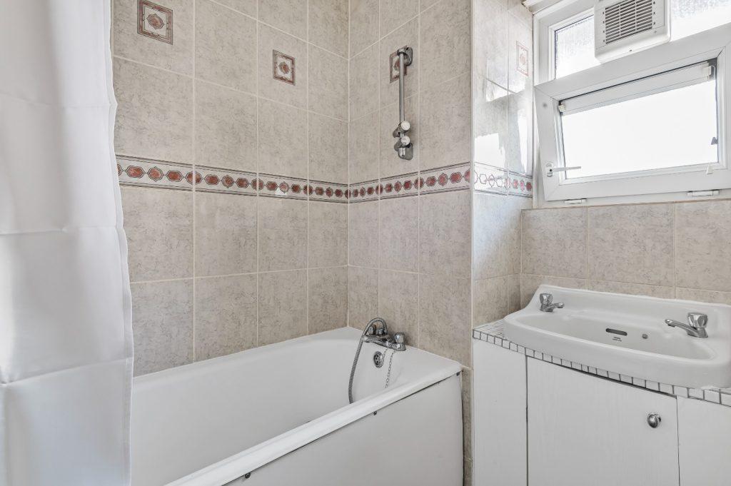 A bathroom in Flat 3 with beige tiled walls and a white bathtub. Above the bathtub is a showerhead and faucets, with a curtain on one side. A small sink with a cabinet sits below an open window, making it an attractive feature of Macclesfield House Central Street LONDON EC1V 8BA investment for sale by auction. Presented by Palace Auctions