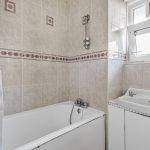 A bathroom in Flat 3 with beige tiled walls and a white bathtub. Above the bathtub is a showerhead and faucets, with a curtain on one side. A small sink with a cabinet sits below an open window, making it an attractive feature of Macclesfield House Central Street LONDON EC1V 8BA investment for sale by auction. Presented by Palace Auctions