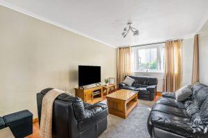 A cozy living room in Flat 3, Macclesfield House, Central Street LONDON EC1V 8BA features black leather sofas arranged around a wooden coffee table. A flatscreen TV sits on a wooden stand against the wall, with light-colored curtains framing a window that offers a view of nearby buildings. A small black stool is in the corner. Presented by Palace Auctions