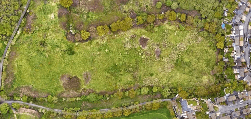 Property for sale by Auction Aerial view of a large open green field with patches of trees and shrubs along the perimeter. A curved road runs along the bottom and right edges of the image. Houses are clustered in the bottom right corner near the road. Presented by Palace Auctions