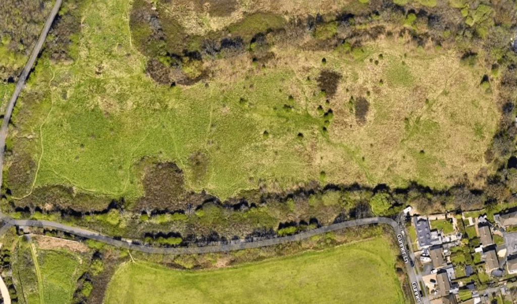 Property for sale by Auction Aerial view of a landscape featuring a large, open grassy area with patches of trees and shrubs. A narrow road runs diagonally near the bottom, with a few buildings and houses clustered on the right side. Pathways meander through the greenery on the left. Presented by Palace Auctions