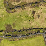 Property for sale by Auction Aerial view of a landscape featuring a large, open grassy area with patches of trees and shrubs. A narrow road runs diagonally near the bottom, with a few buildings and houses clustered on the right side. Pathways meander through the greenery on the left. Presented by Palace Auctions