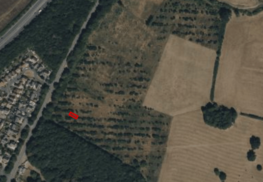 Aerial view of a landscape with patches of forest and open fields. A small, red rectangular highlight marks a specific location within the wooded area. The left side of the image shows a residential neighborhood with a network of roads and houses. Presented by Palace Auctions