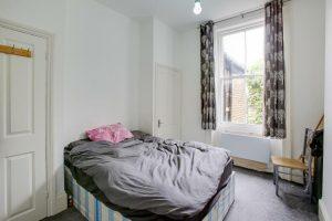 A small bedroom at 33 Spencer Road, Flat 2, featuring a neatly made bed with grey bedding and pink pillows, placed against a white wall. A large window with patterned curtains lets in natural light, while a white door is on the left side of the room and a chair is positioned against the right wall.