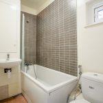 A small, white bathroom in a flat located at 33 Spencer Road, London, W3 6DW, features a bathtub with a gray tiled backsplash, a wall-mounted sink above a small cabinet, and a toilet. A small window on the right side of the wall allows natural light into the space.