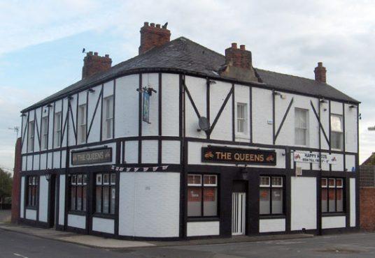 A two-story, white building with black timber accents and red brick chimneys has signs reading "The Queens" and "Happy Halloween" across its facade. It stands at a street corner and has multiple windows and a red and white door. Presented by Palace Auctions