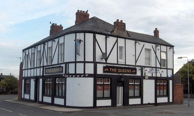 A two-story, white building with black timber accents and red brick chimneys has signs reading "The Queens" and "Happy Halloween" across its facade. It stands at a street corner and has multiple windows and a red and white door. Presented by Palace Auctions