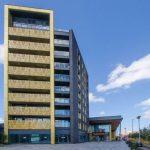 A beautiful modern London apartment, available for sale by auction with Palace Auctions London, stands under a partly cloudy blue sky. The building features a stylish mix of yellow and black exterior finishes, with a Sainsbury's store entrance on the right. In the foreground, there's a sidewalk and paved area adorned with plants.