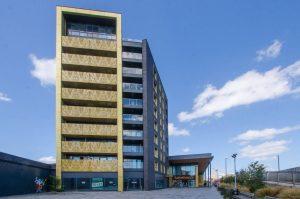 A beautiful modern London apartment, available for sale by auction with Palace Auctions London, stands under a partly cloudy blue sky. The building features a stylish mix of yellow and black exterior finishes, with a Sainsbury's store entrance on the right. In the foreground, there's a sidewalk and paved area adorned with plants.