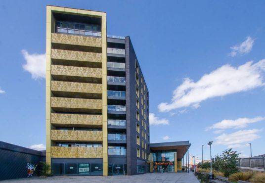 A beautiful modern London apartment, available for sale by auction with Palace Auctions London, stands under a partly cloudy blue sky. The building features a stylish mix of yellow and black exterior finishes, with a Sainsbury's store entrance on the right. In the foreground, there's a sidewalk and paved area adorned with plants.
