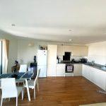 A beautiful modern London apartment available for sale by auction with Palace Auctions London features a minimalist kitchen with white cabinets, a white refrigerator, and a black oven. A glass dining table with white chairs sits on a wooden floor, while sliding glass doors lead to a brightly lit balcony.