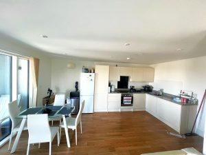 A beautiful modern London apartment available for sale by auction with Palace Auctions London features a minimalist kitchen with white cabinets, a white refrigerator, and a black oven. A glass dining table with white chairs sits on a wooden floor, while sliding glass doors lead to a brightly lit balcony.