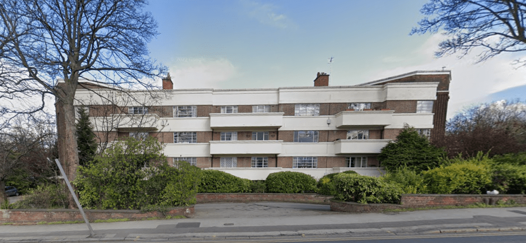 The three-story residential building at 29 Mansfield Court, Nottingham, features a mix of red brick and white concrete facade. Each apartment has a balcony, surrounded by trees and shrubbery. A paved driveway and sidewalk are visible in the foreground with a blue sky above. NG5 2BW for sale by auction with Palace Auctions.