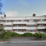The three-story residential building at 29 Mansfield Court, Nottingham, features a mix of red brick and white concrete facade. Each apartment has a balcony, surrounded by trees and shrubbery. A paved driveway and sidewalk are visible in the foreground with a blue sky above. NG5 2BW for sale by auction with Palace Auctions.