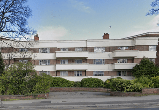 The three-story residential building at 29 Mansfield Court, Nottingham, features a mix of red brick and white concrete facade. Each apartment has a balcony, surrounded by trees and shrubbery. A paved driveway and sidewalk are visible in the foreground with a blue sky above. NG5 2BW for sale by auction with Palace Auctions.