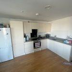 A beautiful modern London apartment available for sale by auction with Palace Auctions London features a modern kitchen with white cabinets and a hardwood floor. The appliances include a white refrigerator, an oven with an electronic stovetop, and a microwave. Black countertops hold various supplies and small kitchen appliances.