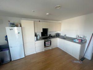 A beautiful modern London apartment available for sale by auction with Palace Auctions London features a modern kitchen with white cabinets and a hardwood floor. The appliances include a white refrigerator, an oven with an electronic stovetop, and a microwave. Black countertops hold various supplies and small kitchen appliances.