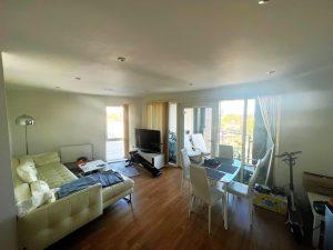 A beautiful modern London apartment available for sale by auction through Palace Auctions London features a bright living room with light-colored walls and wooden flooring. It has a white sectional sofa, a TV stand, an exercise bike, a dining table with four chairs, and large windows letting in plenty of daylight. Various items are scattered around.