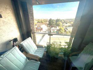 A cozy balcony with two cushioned chairs and several plants in pots. The balcony has a glass and metal railing with a protective net. The background features a view of a park, buildings, and greenery under a clear sky. This beautiful modern London apartment is available for sale by auction with Palace Auctions London.