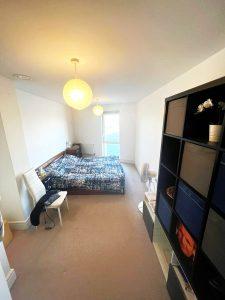A cozy bedroom in a beautiful modern London apartment features a bed with a blue and white patterned duvet, a white chair with a black jacket draped over it, a black shelving unit, and a tall white cabinet. A glowing pendant lamp hangs from the ceiling, while the window with balcony door illuminates the space. This stunning property is available for sale by auction with Palace Auctions and Palace Auctions London