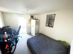 A beautiful modern London apartment bedroom features a cozy bed with gray bedding, a wardrobe, and a bicycle. A desk with various items sits below a large window adorned with a poster. Lit by natural and ceiling light, this stunning space is available for sale by auction with Palace Auctions London.