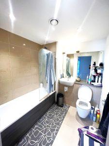 A beautiful modern London apartment available for sale by auction features a bathroom with tiled walls and floors, a bathtub on the left, and a toilet and sink on the right. Towels hang on a rail over the bathtub, while multiple toiletries are visible on the counter and adjacent shelves.