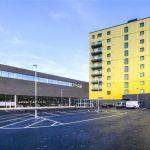 A beautiful modern London apartment is available for sale by auction through Palace Auctions London. This property features a tall, yellow residential tower with multiple balconies on the right side and a low, dark gray commercial structure on the left. A nearly empty parking lot sits in the foreground.