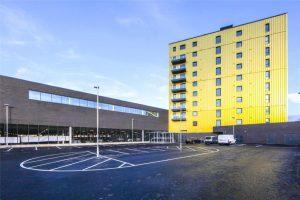 A beautiful modern London apartment is available for sale by auction through Palace Auctions London. This property features a tall, yellow residential tower with multiple balconies on the right side and a low, dark gray commercial structure on the left. A nearly empty parking lot sits in the foreground.