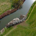 Aerial view of a winding river bordered by lush green fields and a row of flowering trees with white blossoms on 4 acres of land. The landscape, sold by Palace Auctions, is dotted with various shades of green and brown vegetation, creating a serene and picturesque scene.
