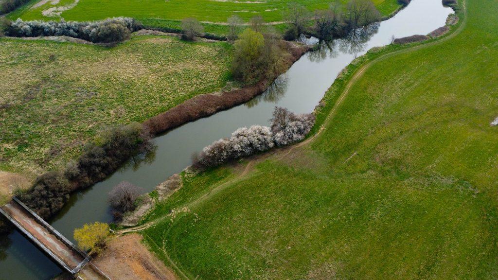 Aerial view of a winding river flowing through lush green fields. A bridge crosses the river on the left side of the image. Trees and shrubs with some blooming white flowers line the riverbank, contrasting with the grassy landscape, within 4 acres of land including access ways sold by Palace Auctions.