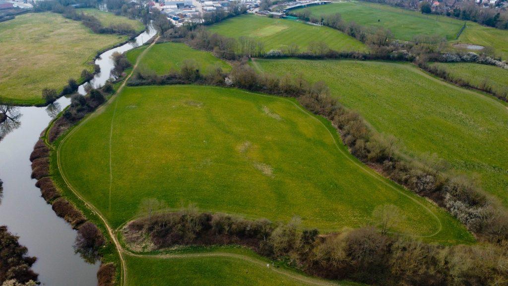 An aerial view of lush, green fields spanning 4 acres, bordered by trees and a winding river. The landscape showcases large open spaces, a few buildings in the background, and patches of bare trees. The river meanders through the land, adding a serene touch to this rural scene sold by Palace Auctions.