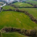 An aerial view of lush, green fields spanning 4 acres, bordered by trees and a winding river. The landscape showcases large open spaces, a few buildings in the background, and patches of bare trees. The river meanders through the land, adding a serene touch to this rural scene sold by Palace Auctions.