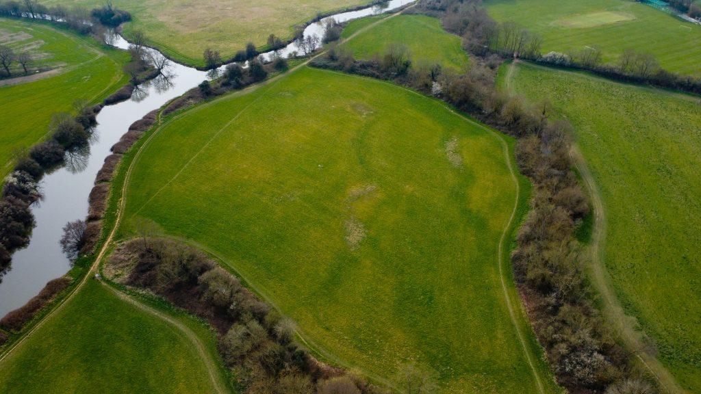 Aerial view of a vibrant green field surrounded by trees and a winding river. The landscape features patches of forested areas and grasslands, separated by natural boundaries. This picturesque 4 acres of land, sold by Palace Auctions, includes access ways, adding to the serene rural scene.