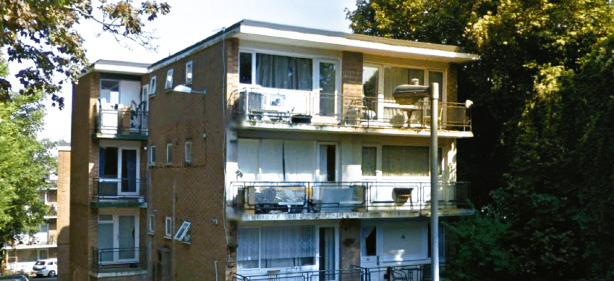 A three-story brick apartment building with multiple balconies and large windows. Some balconies have outdoor furniture. A parking area and several trees are visible in the background. The building is surrounded by greenery.