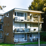 A three-story brick apartment building with multiple balconies and large windows. Some balconies have outdoor furniture. A parking area and several trees are visible in the background. The building is surrounded by greenery.