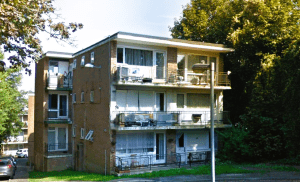 A three-story brick apartment building with multiple balconies and large windows. Some balconies have outdoor furniture. A parking area and several trees are visible in the background. The building is surrounded by greenery.