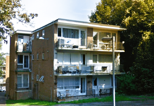 A three-story brick apartment building with multiple balconies and large windows. Some balconies have outdoor furniture. A parking area and several trees are visible in the background. The building is surrounded by greenery.