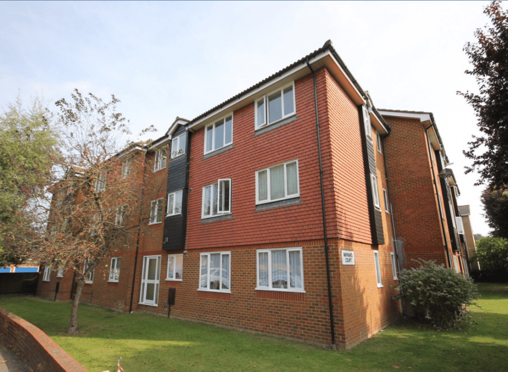 A three-story red brick apartment building with multiple windows and a dark accent panel near the entrance. A small tree, shrubs, and a well-maintained grassy area are in front of the building at 13 Maynard Court, TW18 4QD. Investment apartment available for sale by auction with Palace Auctions. The sky is clear and sunny.