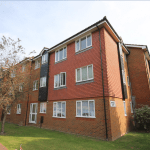 A three-story red brick apartment building with multiple windows and a dark accent panel near the entrance. A small tree, shrubs, and a well-maintained grassy area are in front of the building at 13 Maynard Court, TW18 4QD. Investment apartment available for sale by auction with Palace Auctions. The sky is clear and sunny.