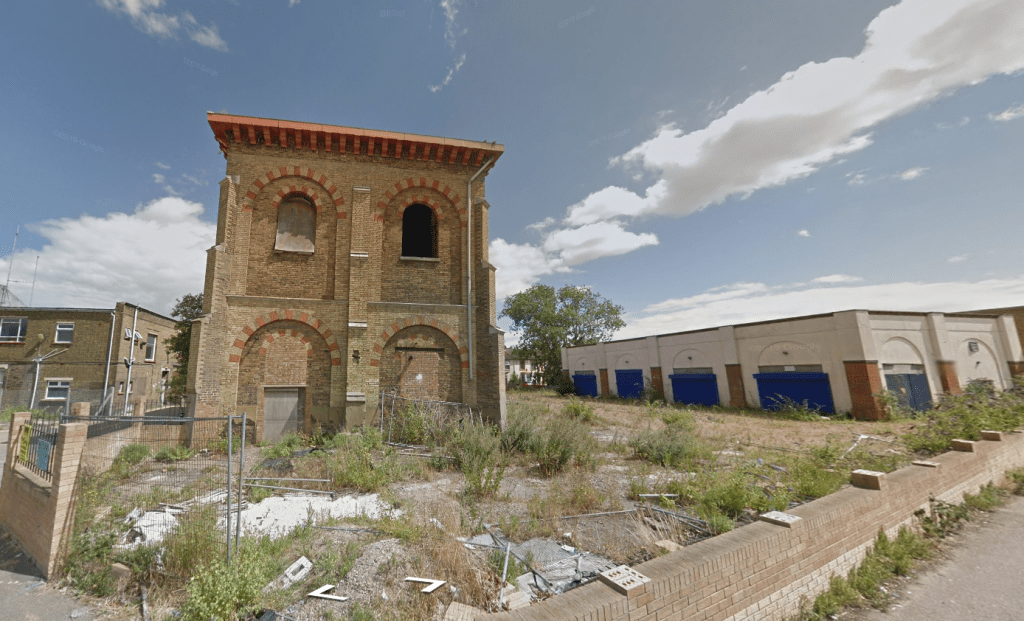 An old, abandoned brick building stands next to a modern, single-story structure with blue shutters. The surrounding area in Kent ME12 2PF is overgrown with weeds, and the sky above is partly cloudy. A brick wall with a metal gate marks the front of this Development Site Sheerness for sale by auction with Palace Auctions.