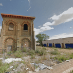An old, abandoned brick building stands next to a modern, single-story structure with blue shutters. The surrounding area in Kent ME12 2PF is overgrown with weeds, and the sky above is partly cloudy. A brick wall with a metal gate marks the front of this Development Site Sheerness for sale by auction with Palace Auctions.