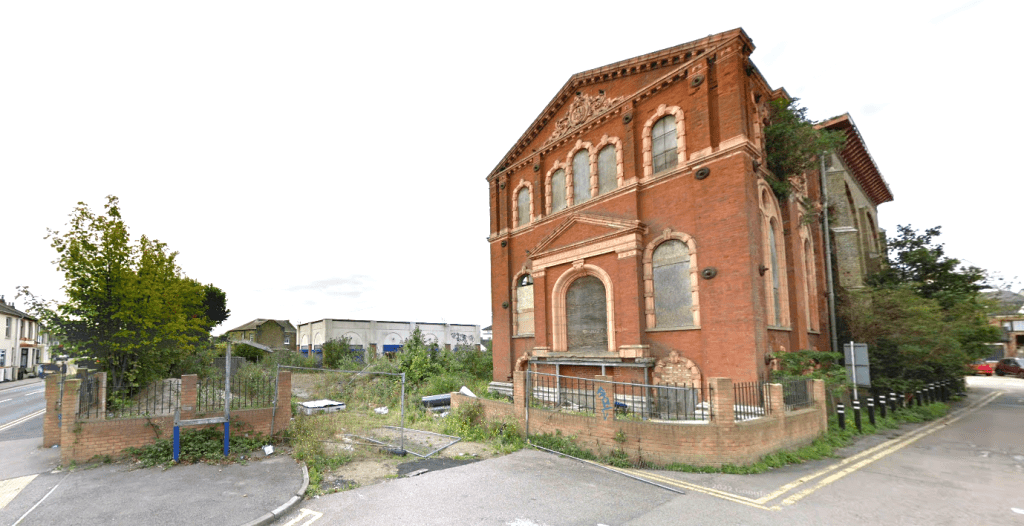 An abandoned, red-brick building with arched windows stands at a street corner in Sheerness, Kent ME12 2PF. Showing signs of decay with broken windows and overgrown vegetation, this development site features a tall, weathered tree on the left and is partially fenced off. For sale by auction with Palace Auctions.