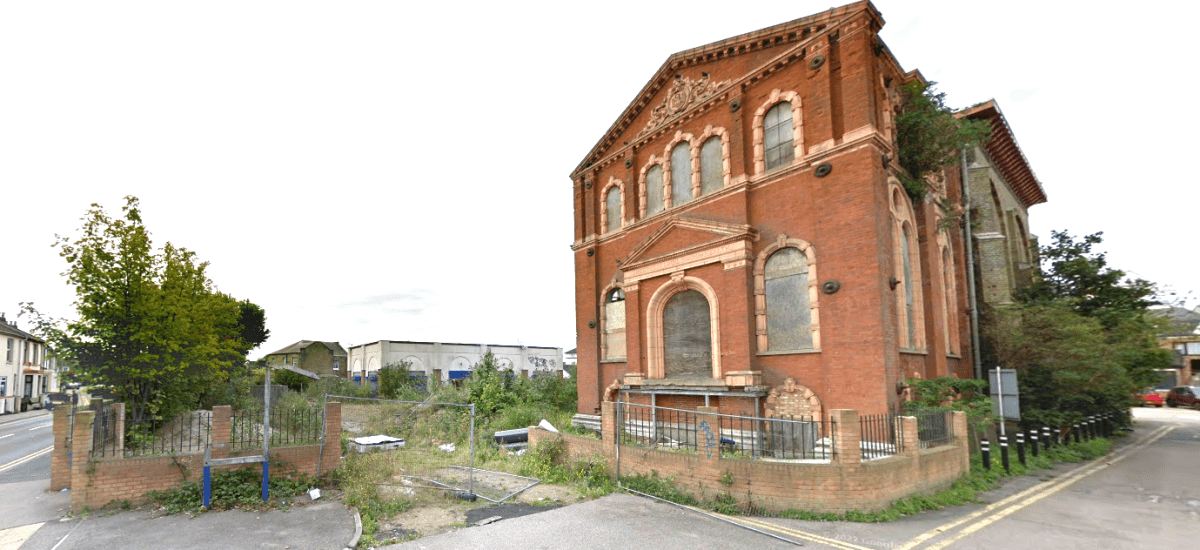 An abandoned, red-brick building with arched windows stands at a street corner in Sheerness, Kent ME12 2PF. Showing signs of decay with broken windows and overgrown vegetation, this development site features a tall, weathered tree on the left and is partially fenced off. For sale by auction with Palace Auctions.