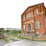 An abandoned, red-brick building with arched windows stands at a street corner in Sheerness, Kent ME12 2PF. Showing signs of decay with broken windows and overgrown vegetation, this development site features a tall, weathered tree on the left and is partially fenced off. For sale by auction with Palace Auctions.