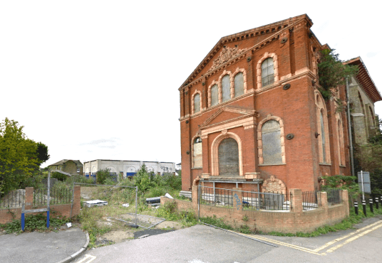 An abandoned, red-brick building with arched windows stands at a street corner in Sheerness, Kent ME12 2PF. Showing signs of decay with broken windows and overgrown vegetation, this development site features a tall, weathered tree on the left and is partially fenced off. For sale by auction with Palace Auctions.