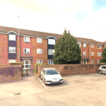 A two-story brick apartment building on Rosefield Road, TW18 4QD, features multiple windows and a tiled roof. There is a small parking area in front with several parked cars. Two large trees are near the entrance, and a brick wall separates the parking lot from the sidewalk. Investment apartment available for sale by auction with Palace Auctions in Staines-upon-Thames.