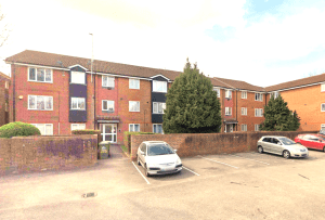 A two-story brick apartment building on Rosefield Road, TW18 4QD, features multiple windows and a tiled roof. There is a small parking area in front with several parked cars. Two large trees are near the entrance, and a brick wall separates the parking lot from the sidewalk. Investment apartment available for sale by auction with Palace Auctions in Staines-upon-Thames.