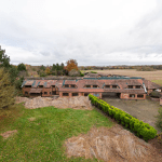 Aerial view of an old brick building with a tiled roof surrounded by overgrown vegetation and fields. The structure shows signs of wear, with some sections of the roof missing. Trees and hedges partially hide parts of the building. Pools Cottages successfully sold at Palace Auctions, revealing an open yard in front.