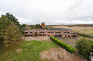 Aerial view of an old brick building with a tiled roof surrounded by overgrown vegetation and fields. The structure shows signs of wear, with some sections of the roof missing. Trees and hedges partially hide parts of the building. Pools Cottages successfully sold at Palace Auctions, revealing an open yard in front.