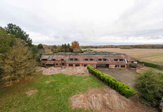 Aerial view of an old brick building with a tiled roof surrounded by overgrown vegetation and fields. The structure shows signs of wear, with some sections of the roof missing. Trees and hedges partially hide parts of the building. Pools Cottages successfully sold at Palace Auctions, revealing an open yard in front.